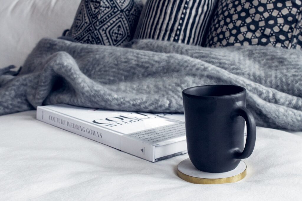 A black ceramic mug is placed on a white and gold coaster beside a book and a gray blanket is on top of a white bed