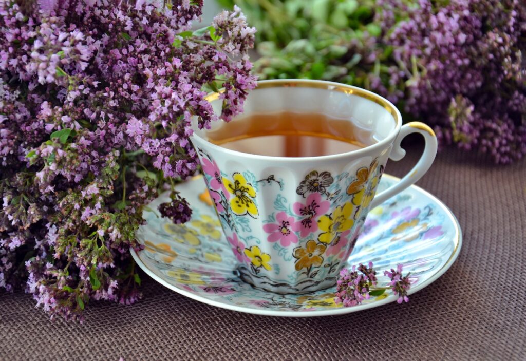 A floral-designed teacup with a matching floral coaster beside purple flowers on top of a brown placemat