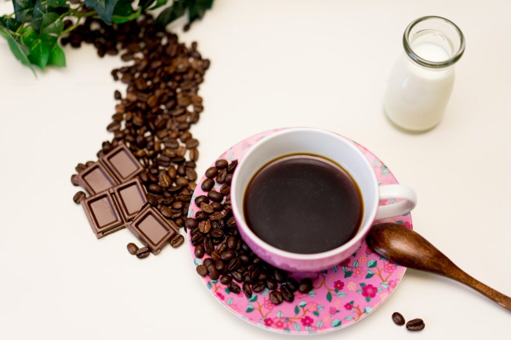 A black coffee on a matching pink floral cup and saucer near five small bars of chocolate, coffee beans, a wooden spoon, and a bottle of milk on a white surface