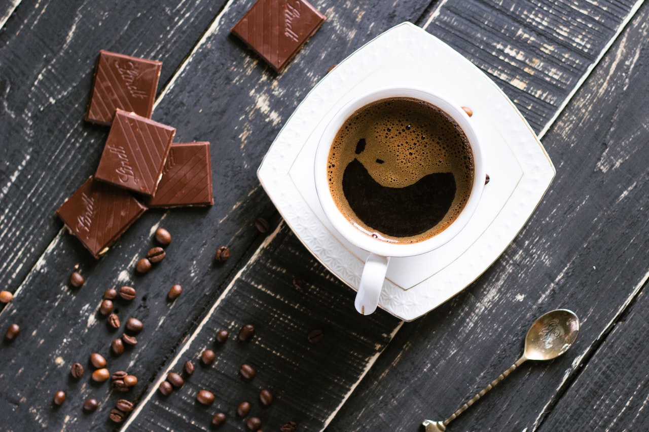 Black coffee in a white cup and saucer near small bars of chocolate, coffee beans, and a golden teaspoon placed on top of a black wooden table
