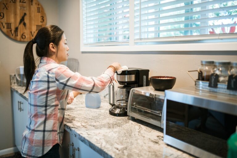 A woman with black hair wearing a checkered long sleeves shirt is wiping a black coffee maker on top of a marbled countertop