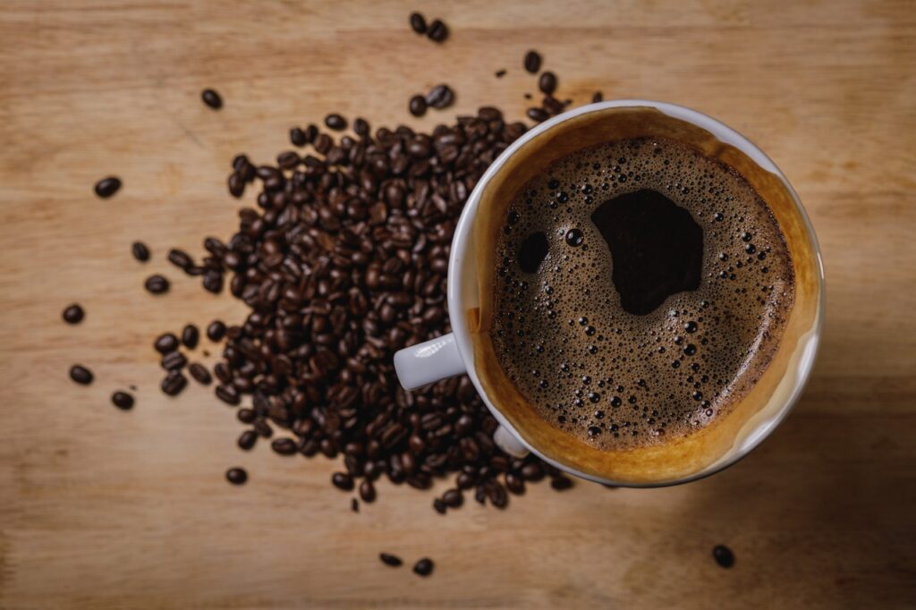 A white cup filled with black coffee beside fresh coffee beans is placed on a brown wooden surface