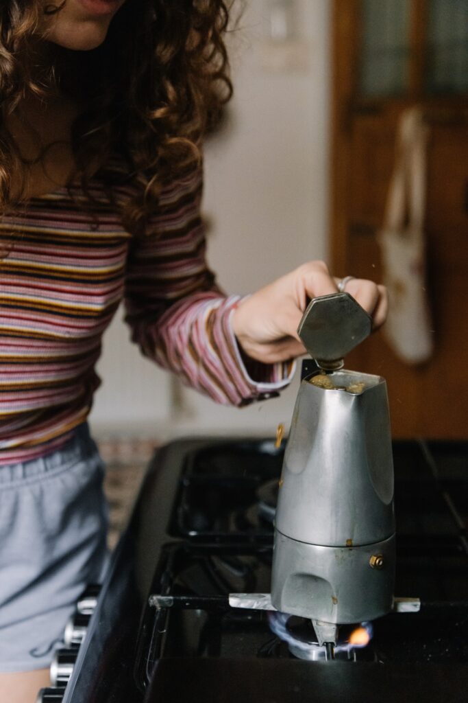 A woman in striped long-sleeves and shorts brewing coffee using a silver Moka pot on a stovetop