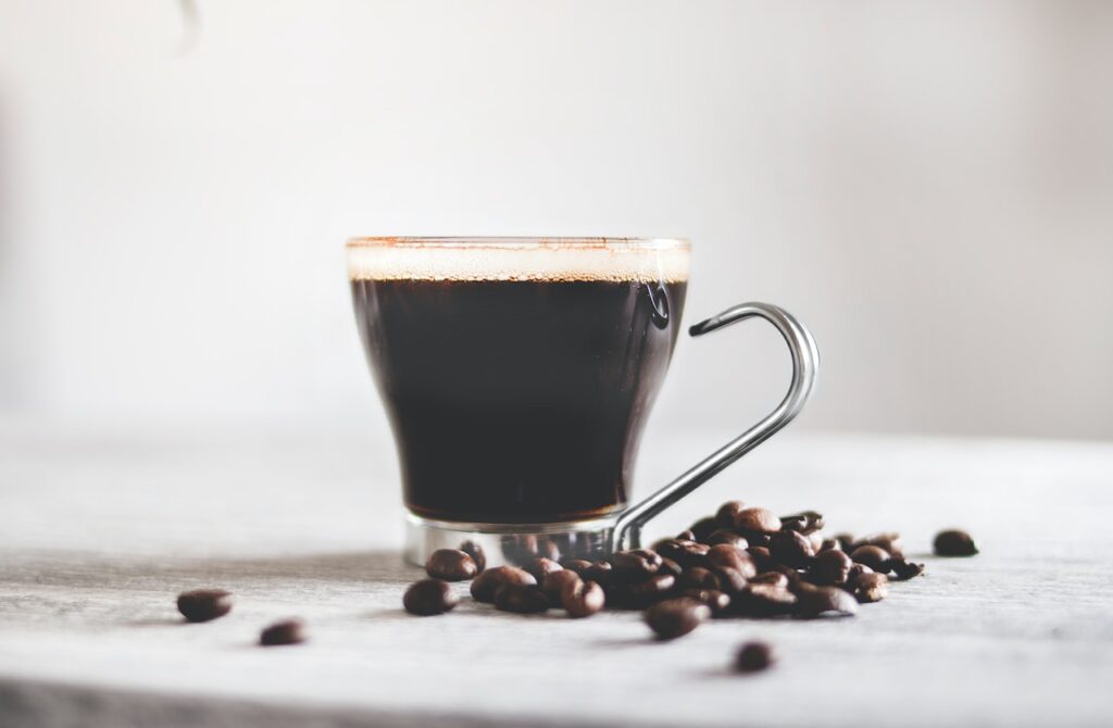 A cup of coffee on a clear small glass beside coffee beans on a white table