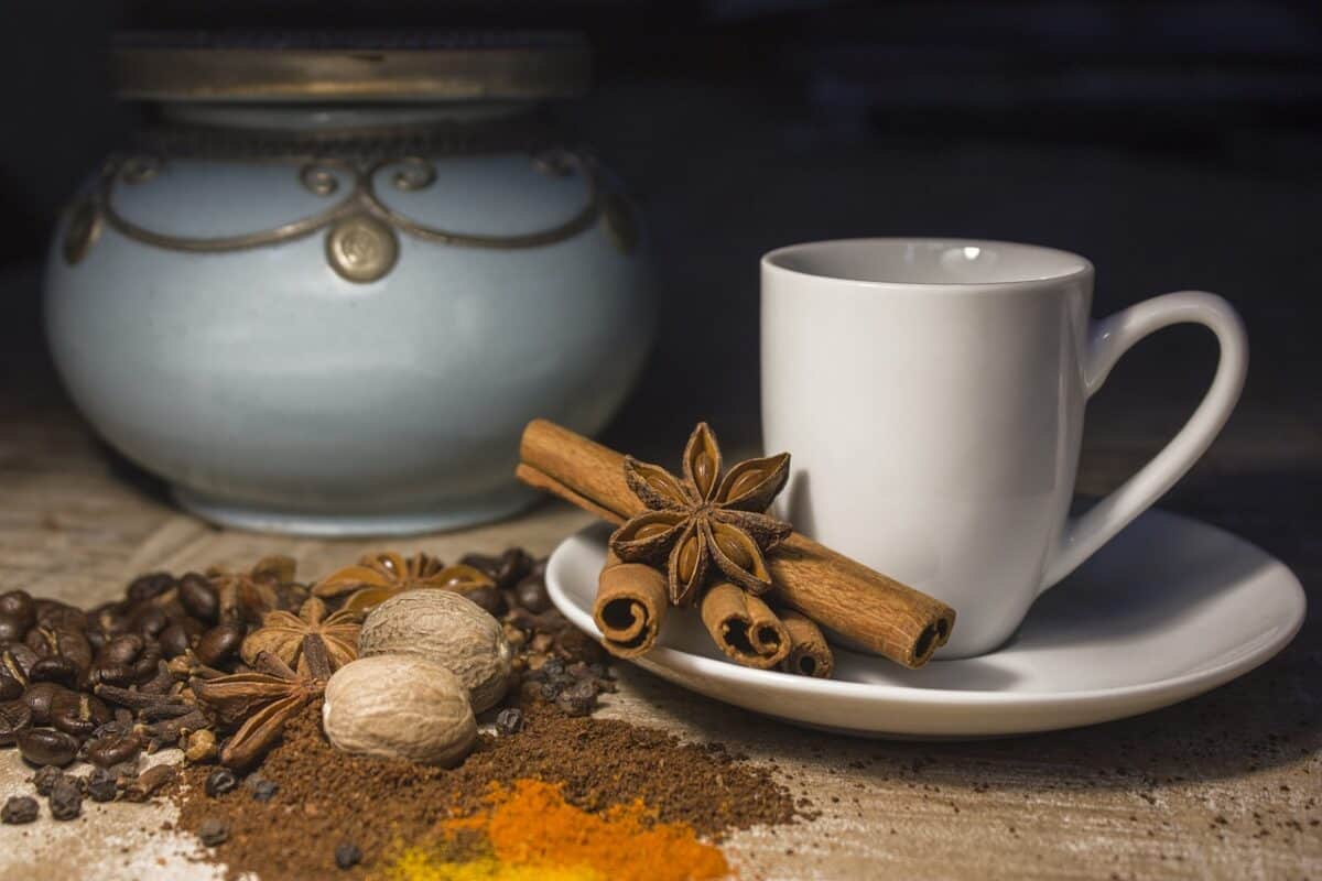 A white mug and cinnamon sticks are placed on a white saucer beside a coffee beans