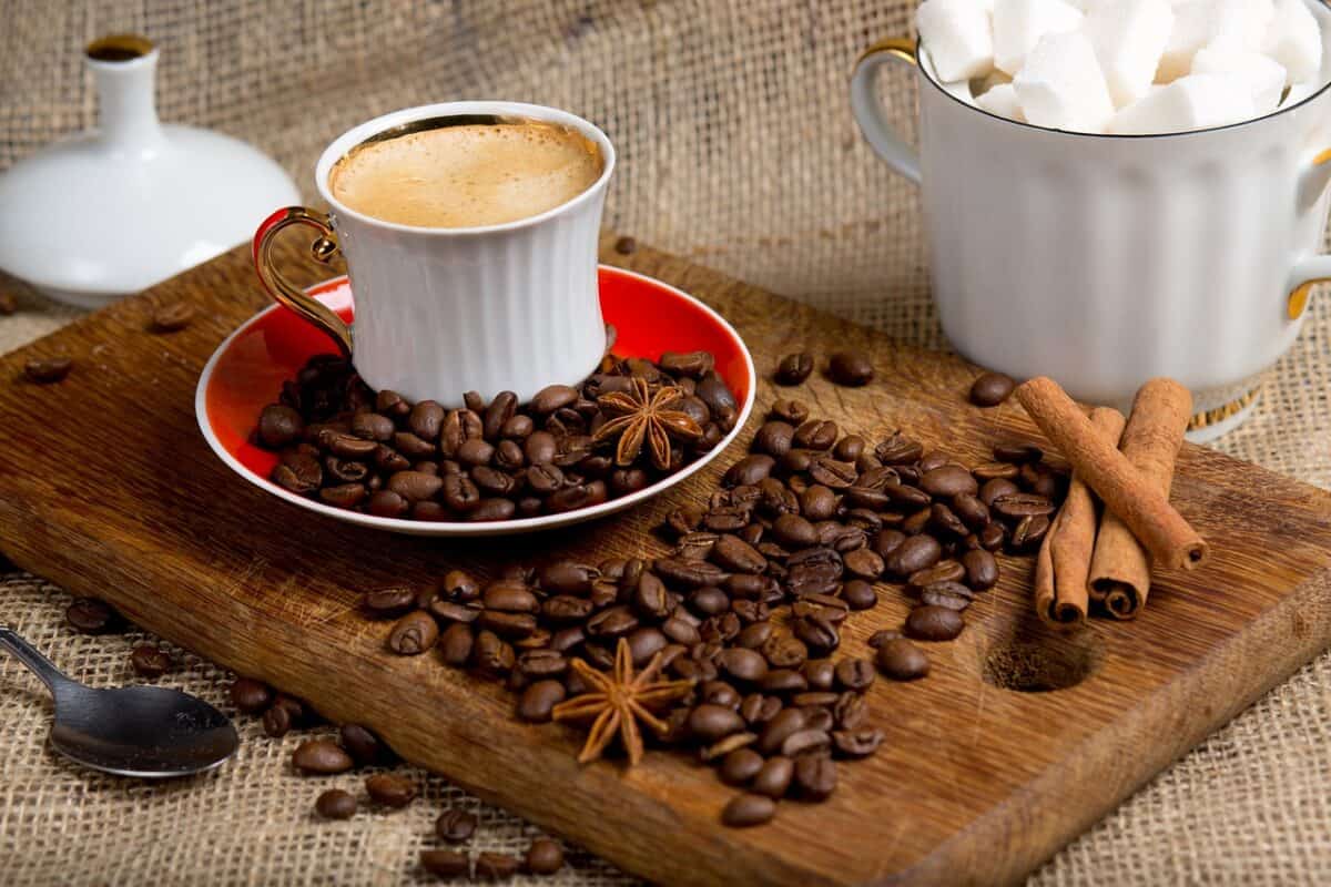 A white mug filled with foamy coffee was placed on a wooden chopping board with coffee beans and cinnamon sticks