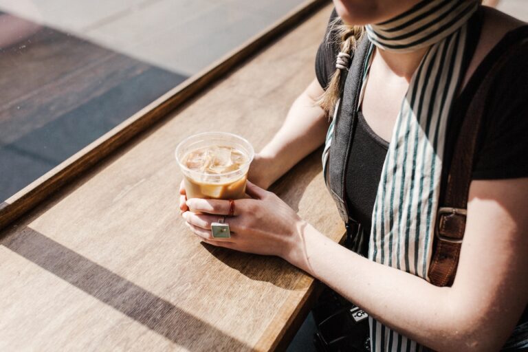 A woman wearing a black blouse and scarf holds an iced coffee placed on a plastic cup