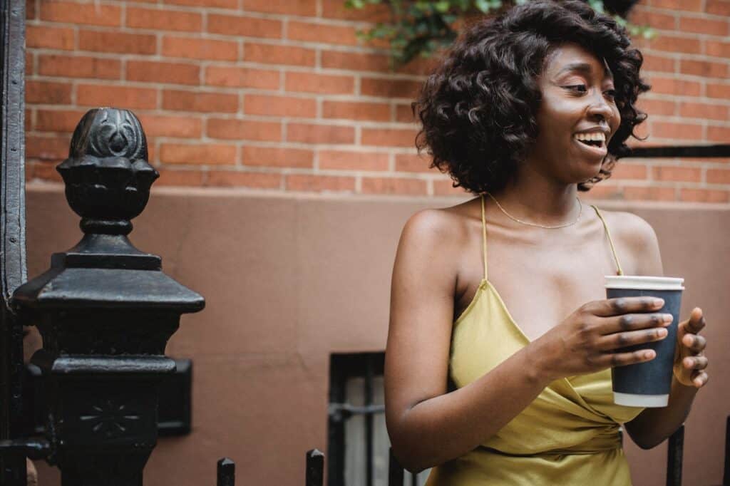 A woman with short curly hair wearing a yellow spaghetti strap dress holds a large cup of coffee outside the apartment