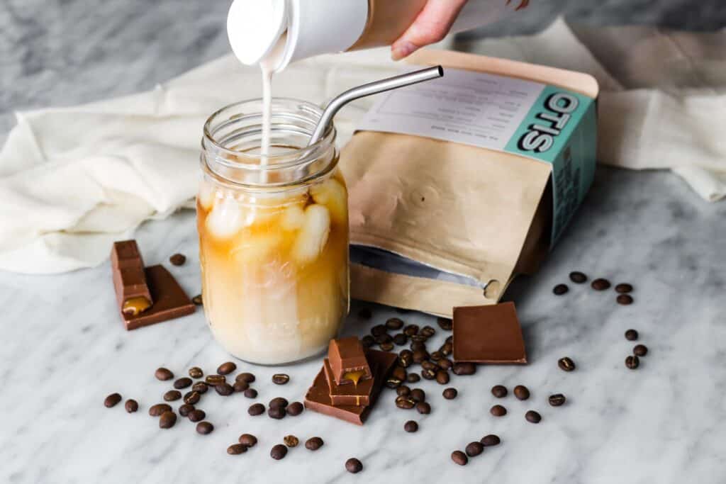 Coffee creamer being poured into a cup of coffee in a mason jar on a quartz countertop surrounded by coffee beans and chocolate