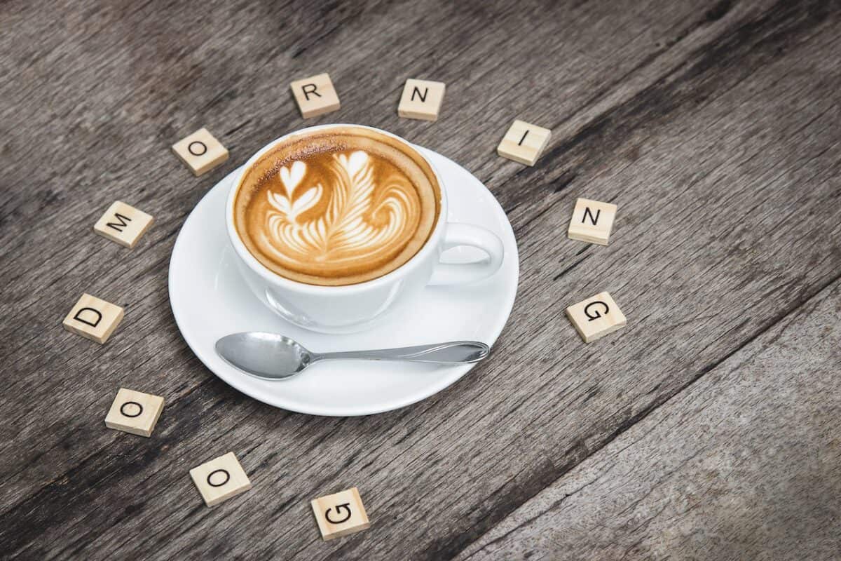 A white ceramic mug with cappuccino coffee beside a silver teaspoon on a white saucer