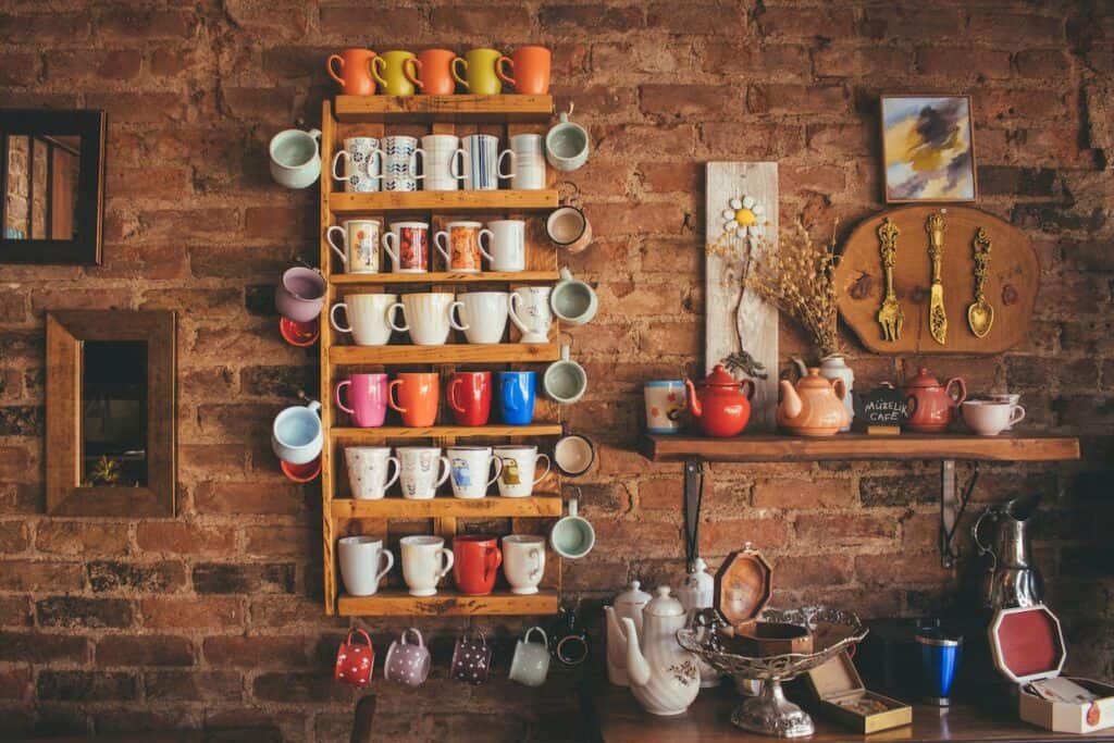 Different colored mugs are arranged on a brown wooden floating rack placed on a brick wall