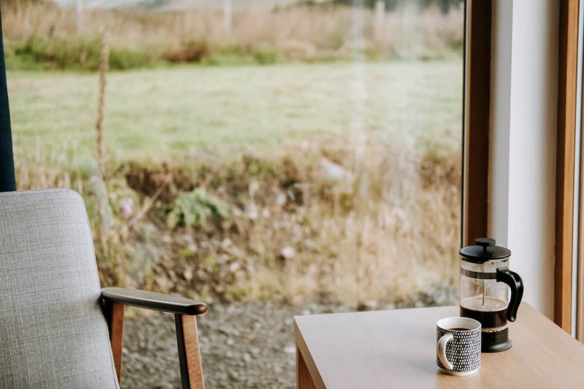 A french press beside a cup of coffee placed on a brown wooden table