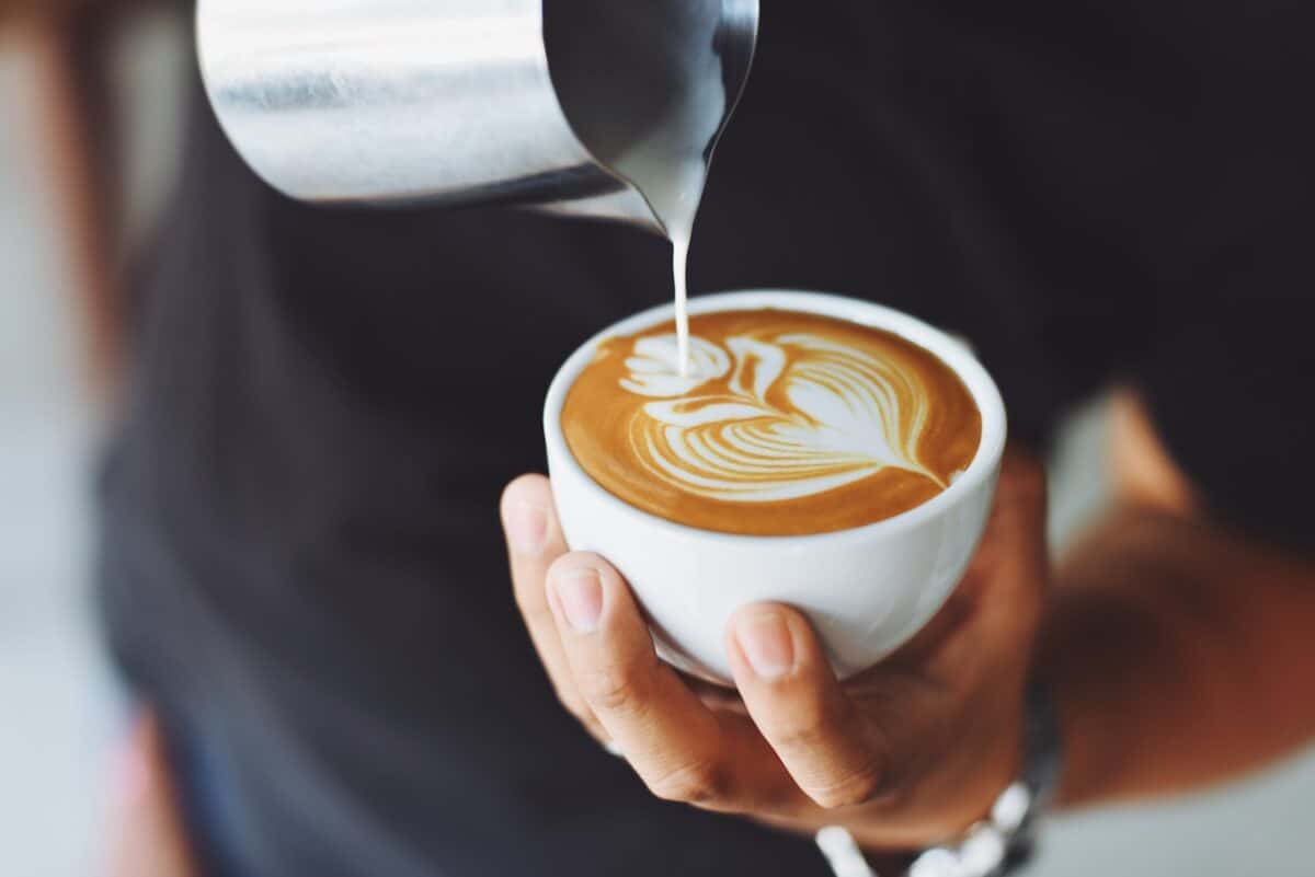 A person wearing a black t-shirt pouring milk placed on stainless steel on a white cup of coffee