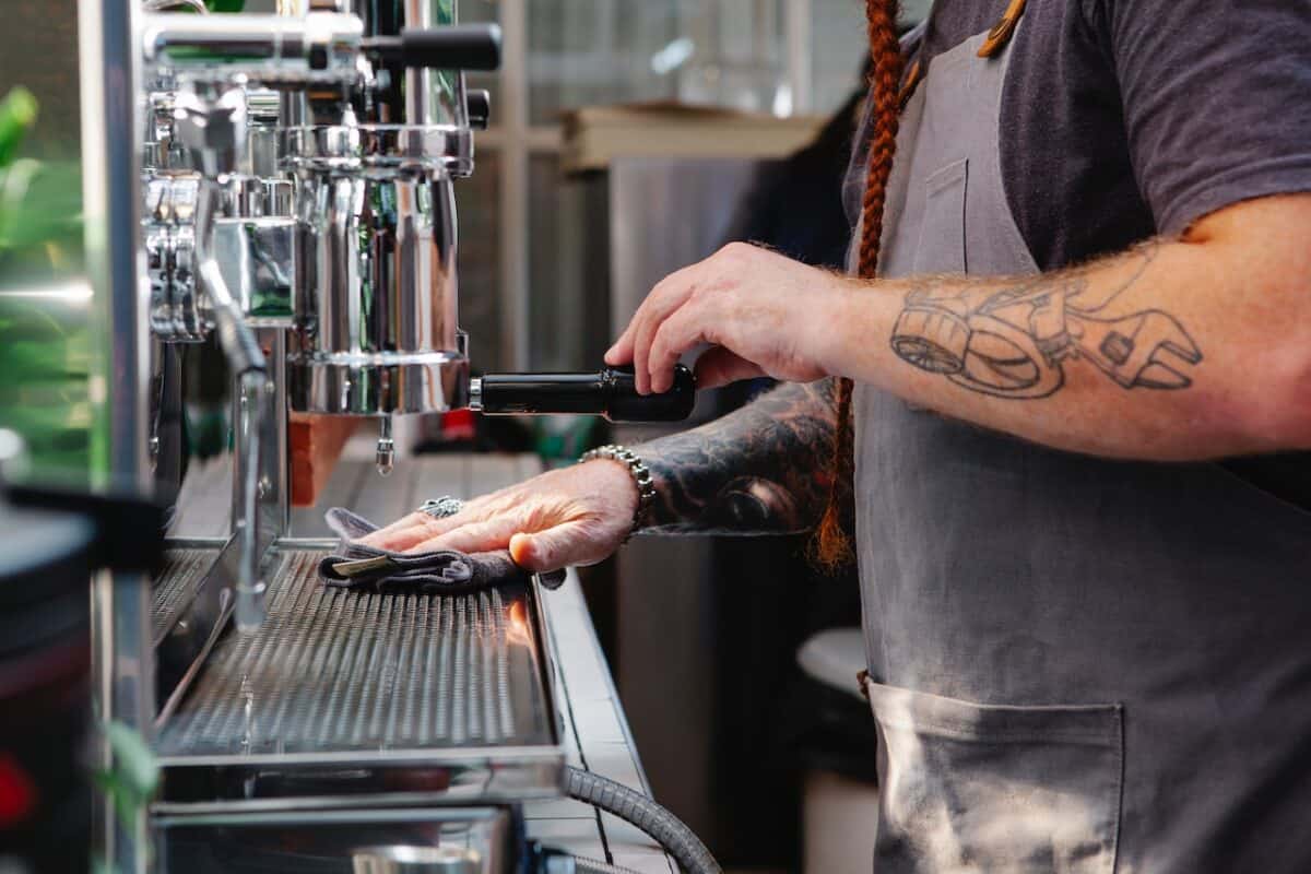 A barista wearing an apron wiping the coffee machine using a rug