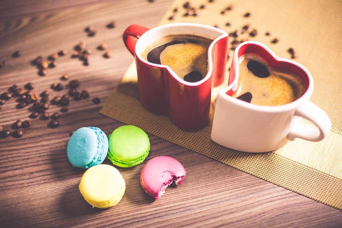 Two heart-shaped cups of coffee beside colorful macaroons and coffee beans