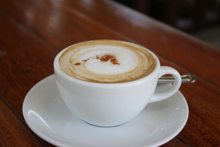 A close-up white cup filled with foamy coffee is placed on a white saucer on a brown wooden table