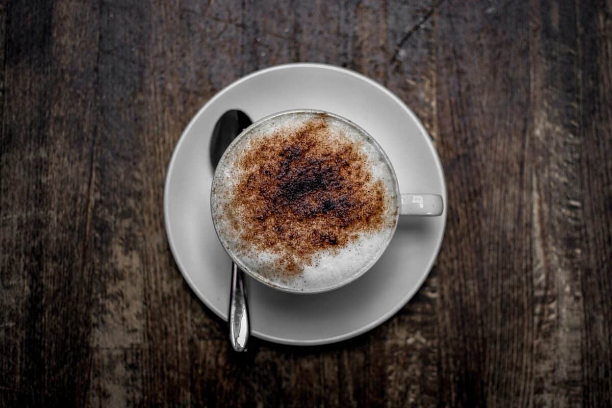 A foamy coffee in a white cup beside a silver spoon is placed on the white saucer on a wooden table