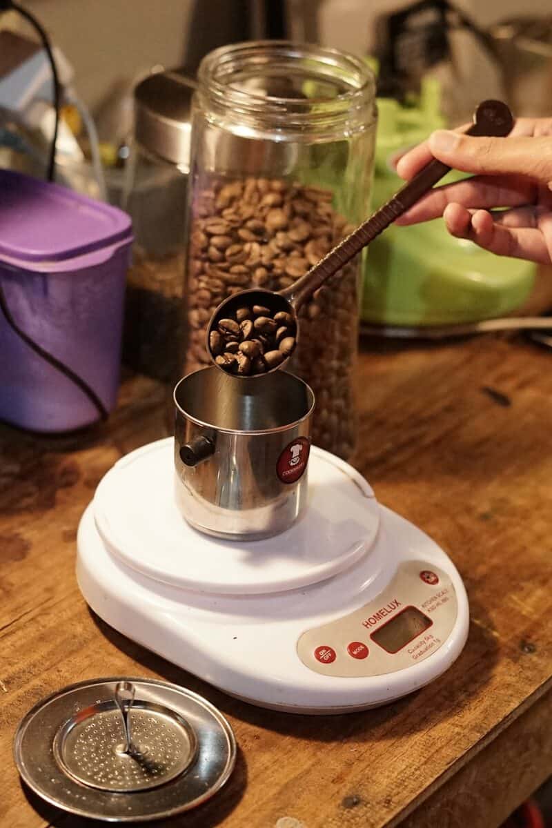 A person pouring coffee beans from a spoon into a small can and weigh it on a white weighing scale