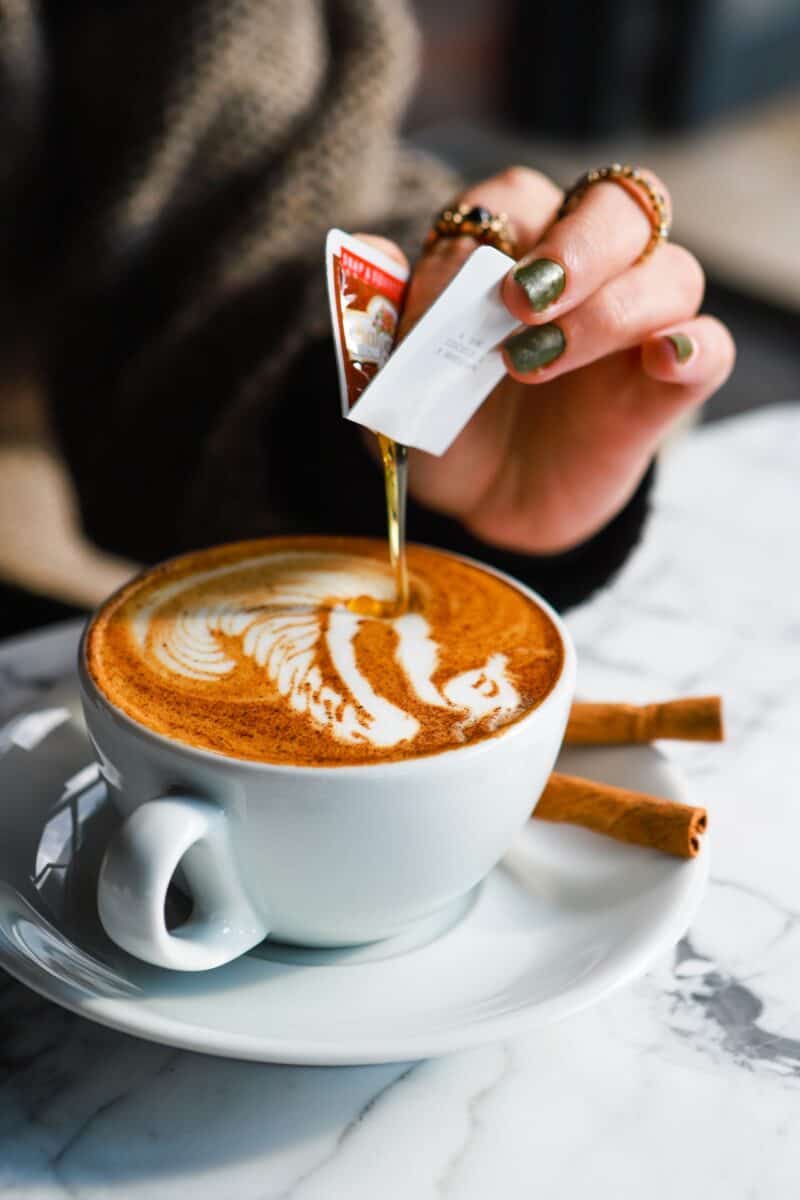 A person with nail polish pouring syrup on a cup of coffee placed on a white saucer