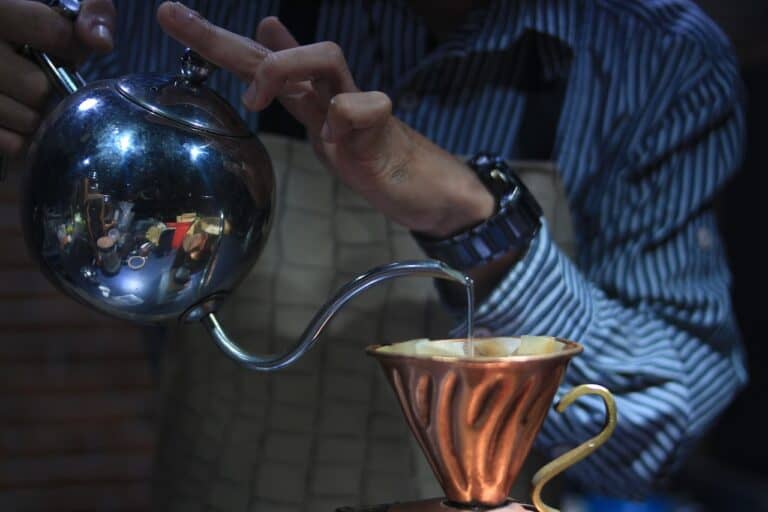 A barista wearing blue striped long sleeves pouring a coffee using a silver kettle