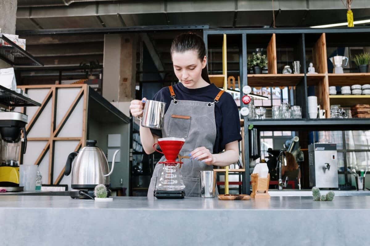A female barista wearing a black t-shirt using a stainless steel mug on pouring water on a pour-over coffee