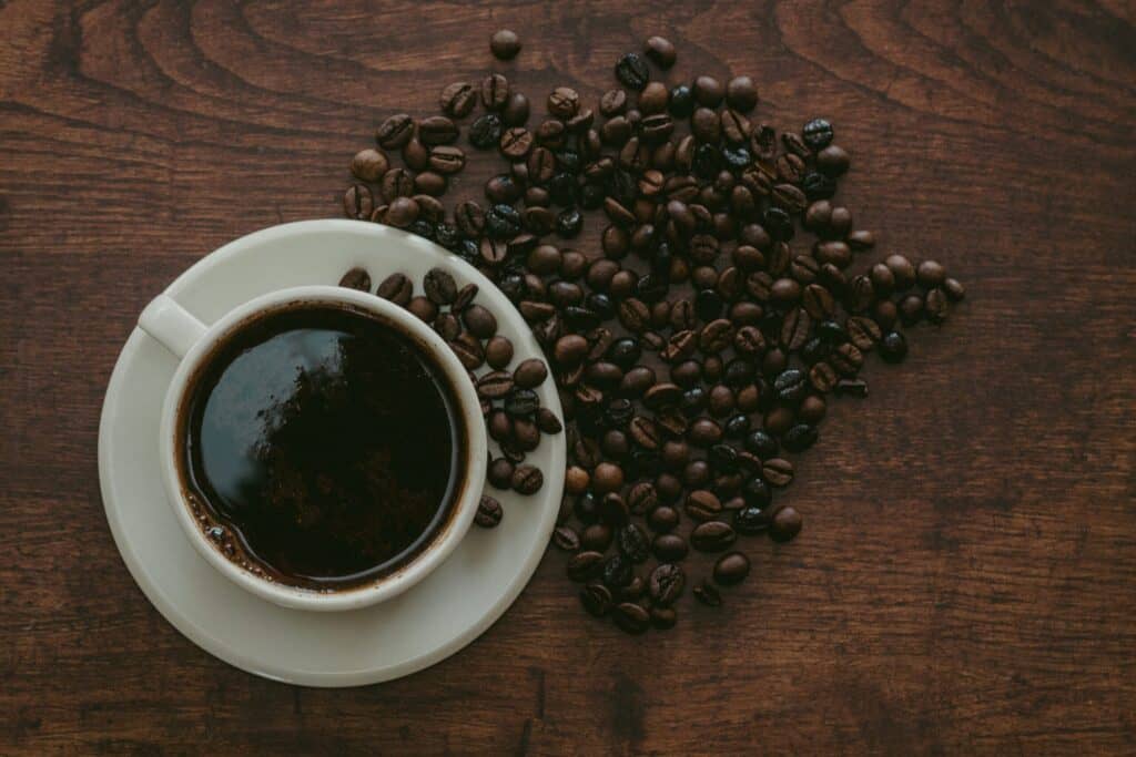 A dark cup of coffee in a white cup and saucer on a brown wooden table with coffee beans spilled on it