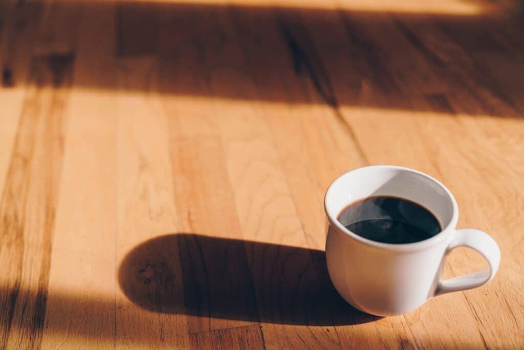 A cup of coffee in a white mug sitting on a light wooden table with the sun shining on it