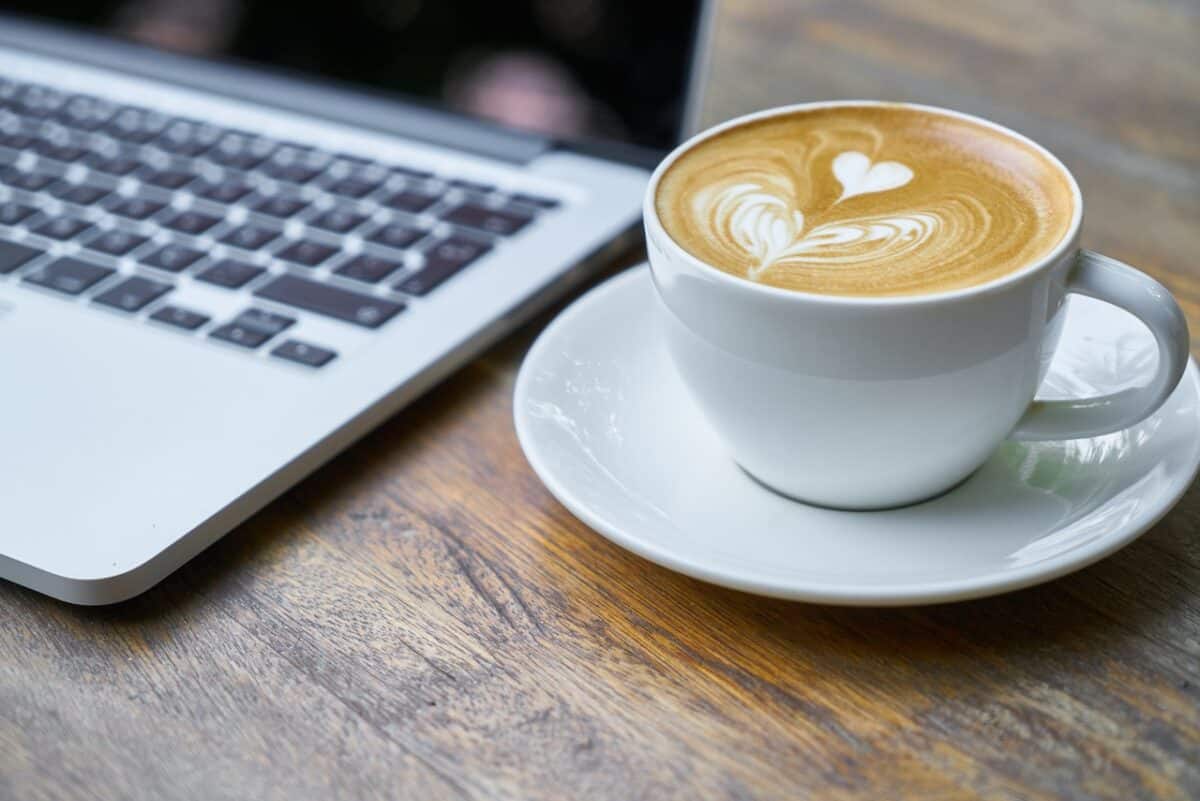 A white cup filled with coffee was placed on a white saucer beside a silver laptop