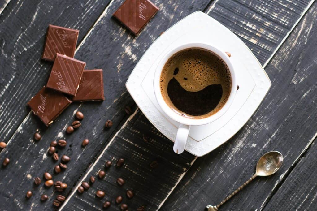 A top view of a white cup of coffee placed on a saucer beside coffee beans and chocolates