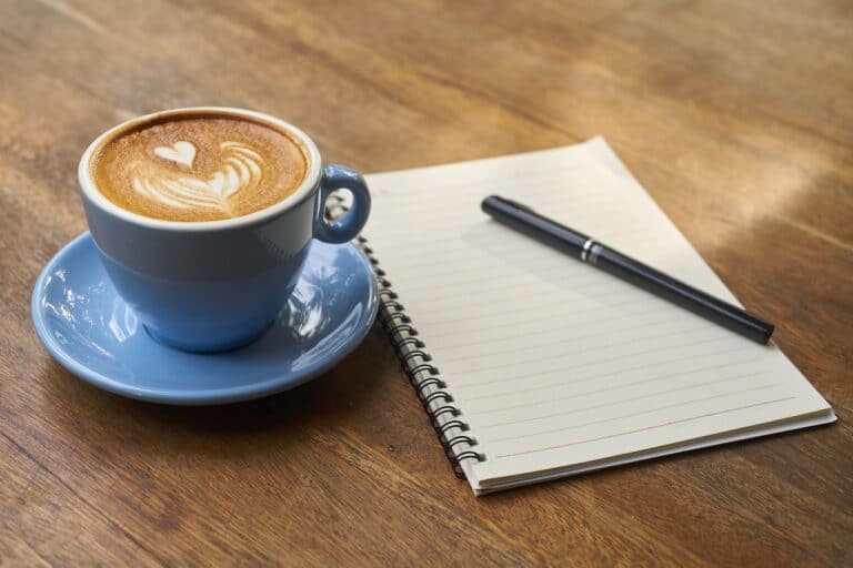 A blue mug filled with coffee was placed on a blue saucer beside a notebook and ballpen