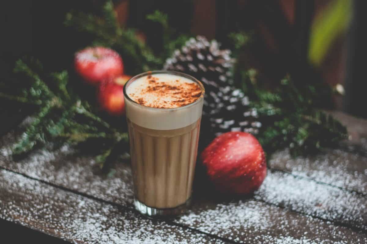 A chai latte sprinkled with cinnamon powder near red apples on top of a brown wood