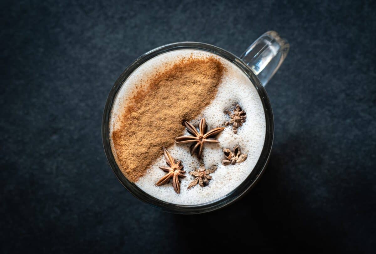Five pieces of star anise and brown cinnamon powder on top of a white coffee foam placed on a clear glass on top of a black surface