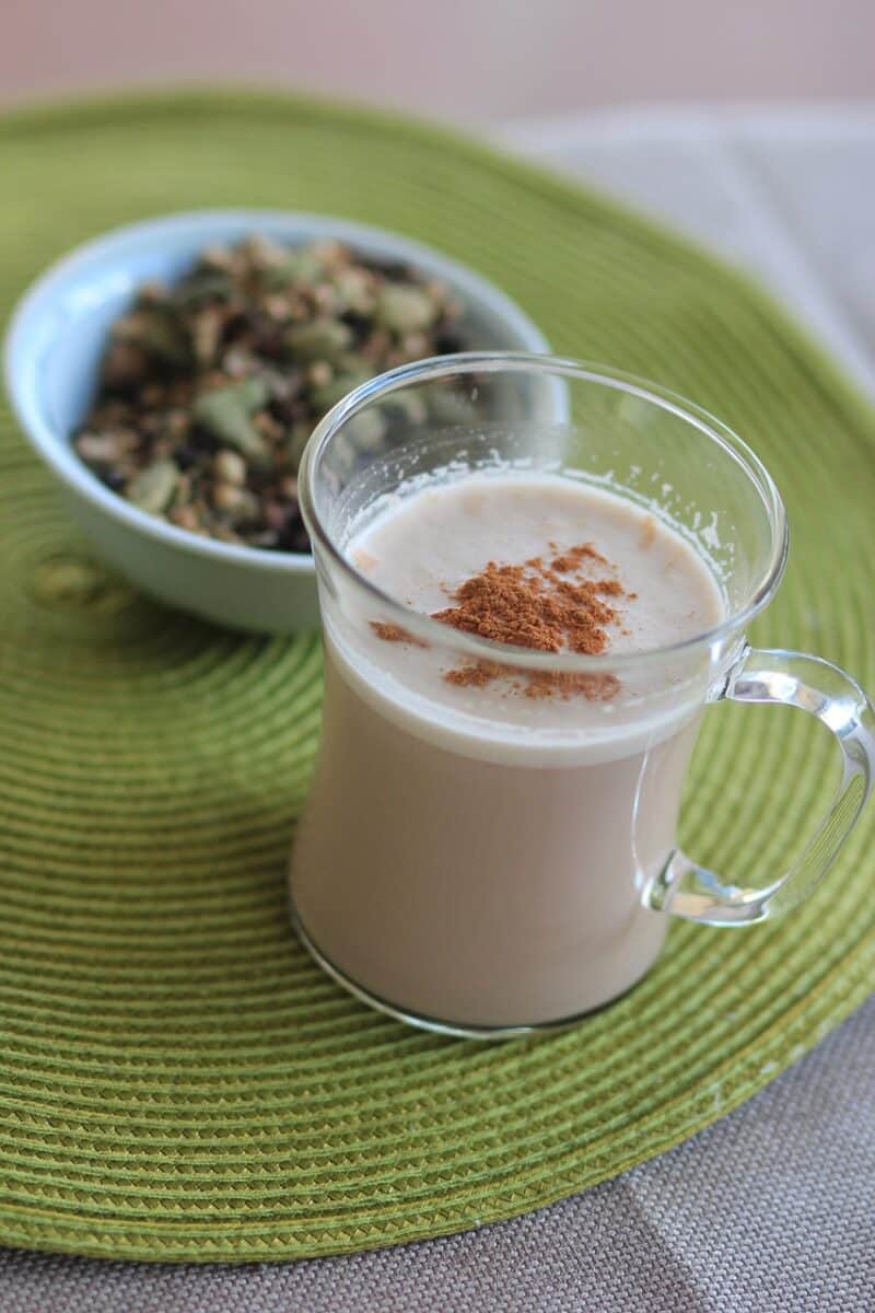 Chai tea with cinnamon sprinkled on a clear glass near a small white bowl on top of a gray placemat