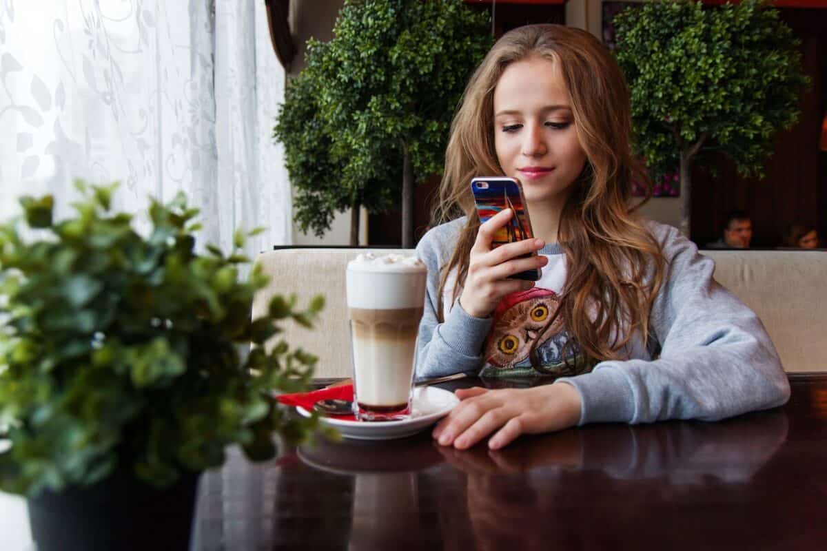 A girl with long brown hair is using her smartphone to take a photo of a glass of latte on a white ceramic saucer on top of a wooden table
