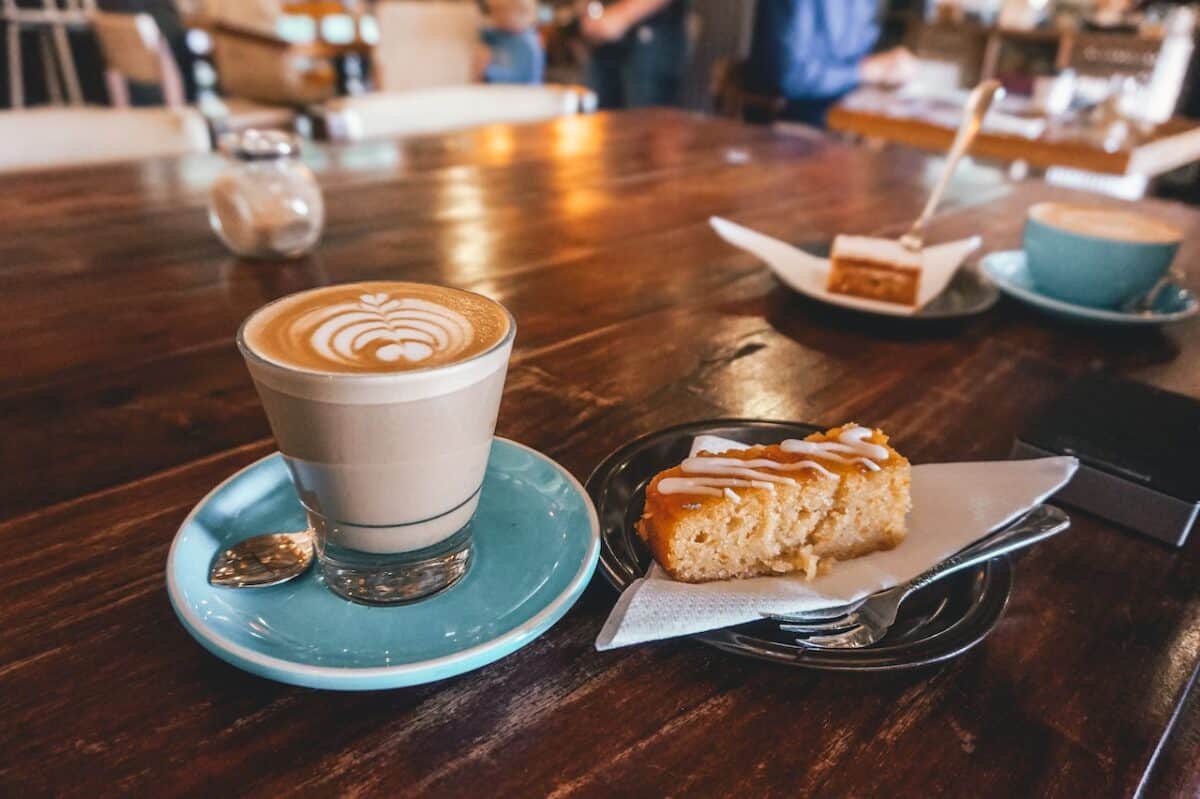 Latte in a clear glass placed on a blue saucer near a slice of cake on top of a wooden table
