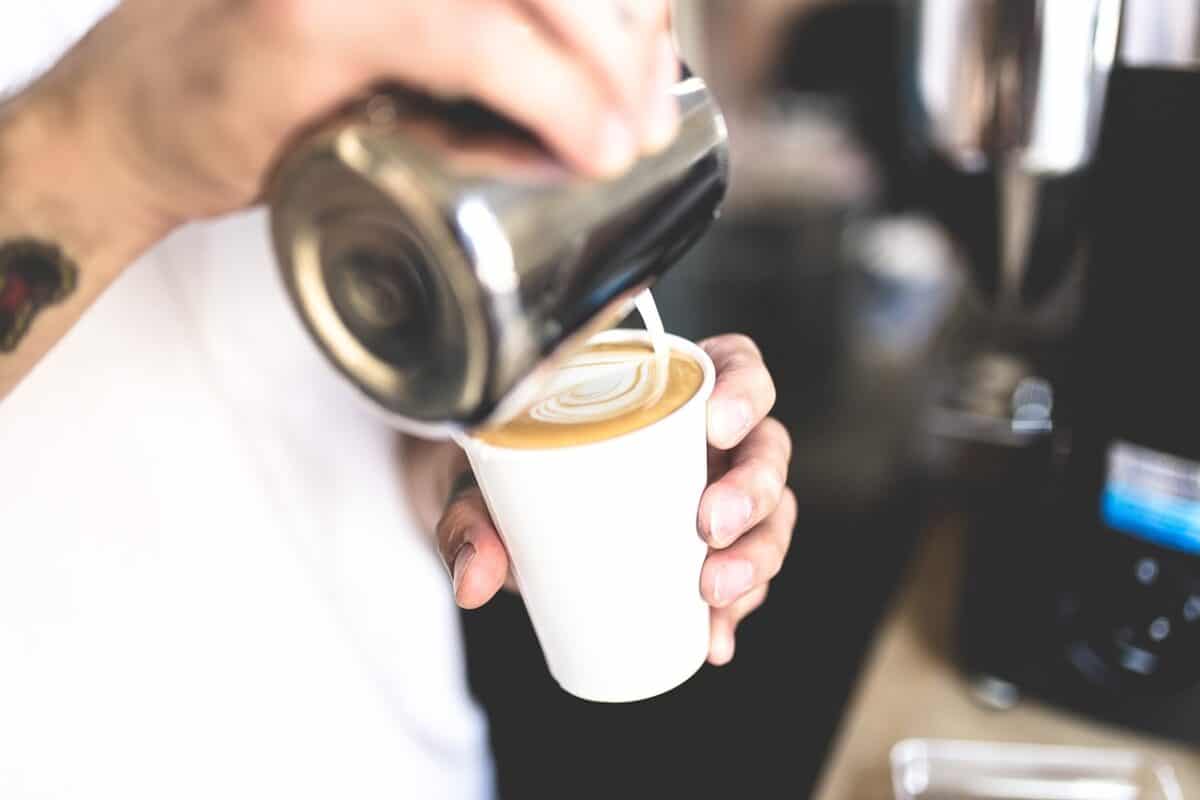 A person wearing a white shirt is pouring milk from a stainless steel cup into a latte on a white cup
