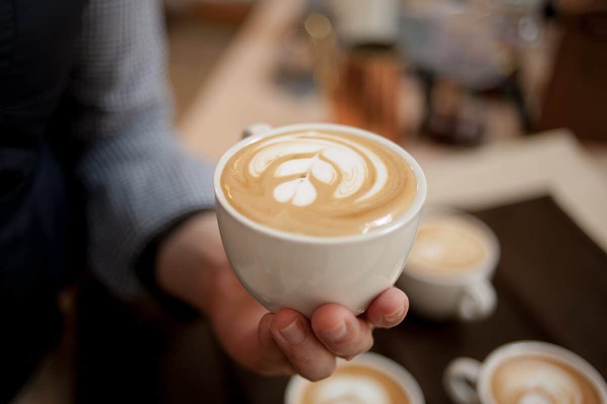 A person wearing a gray long sleeves shirt is holding a white mug filled with latte above three lattes on a white ceramic mug