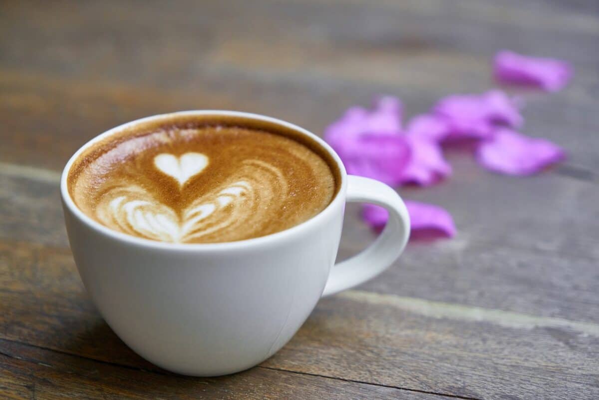 A cup of latte with a white heart coffee design on a white ceramic mug near purple petals and on top of a brown wooden table