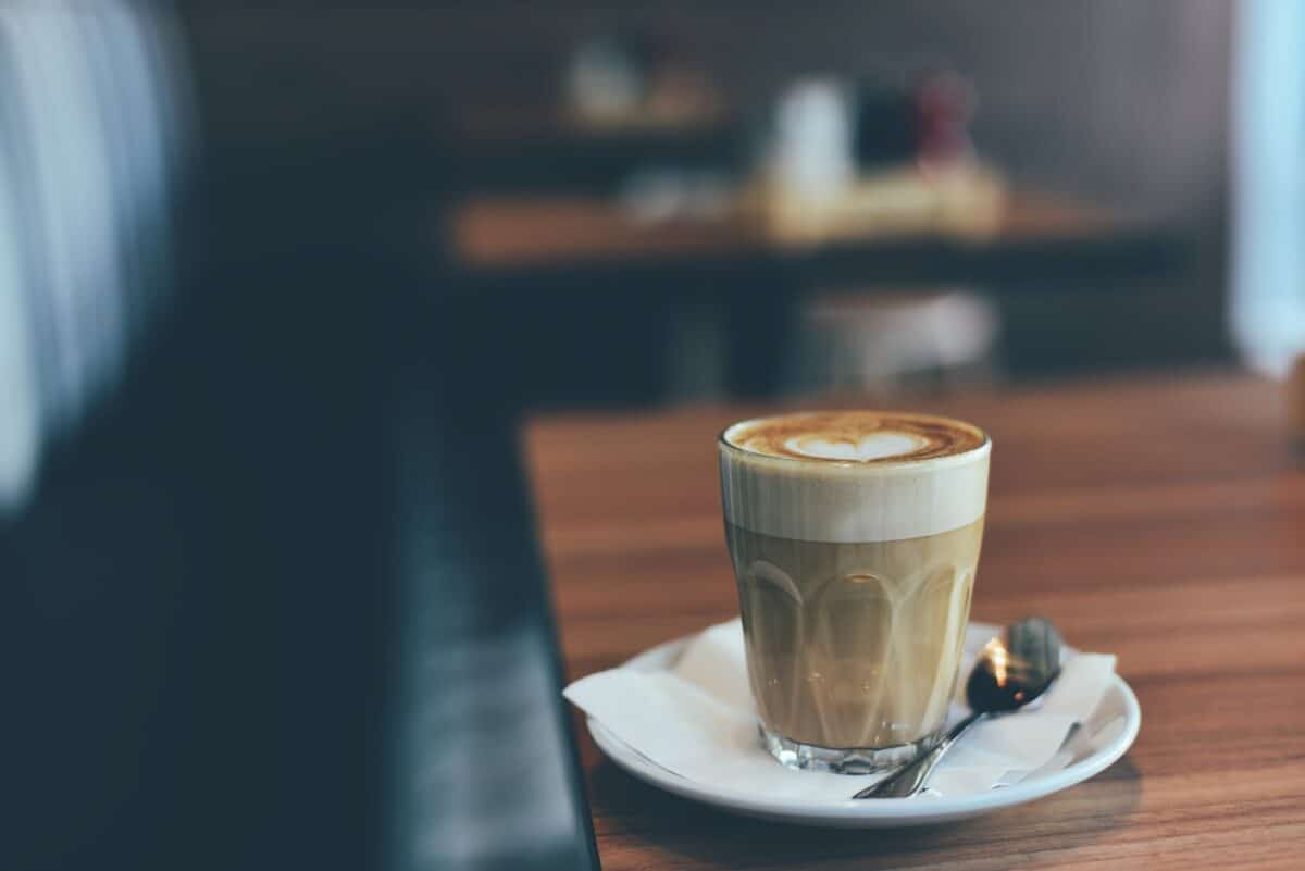 A latte on a clear glass placed on a white saucer on top of a brown wooden table