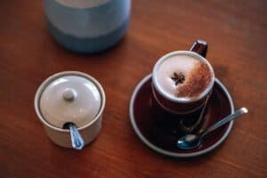 A chai latte on a brown ceramic mug on a brown ceramic saucer near a white ceramic container on top of a brown wooden table